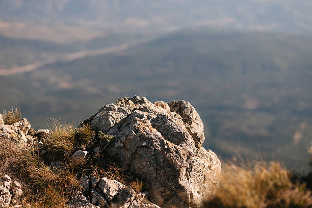 Close up view of a rocky stone with dry grass on mountain top, with blurred valley in the background on a sunny day.