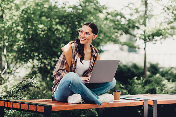 istock Young woman enjoying leisure time outdoors using a laptop, sitting on a bench surrounded by greenery on a sunny day 2231075019
