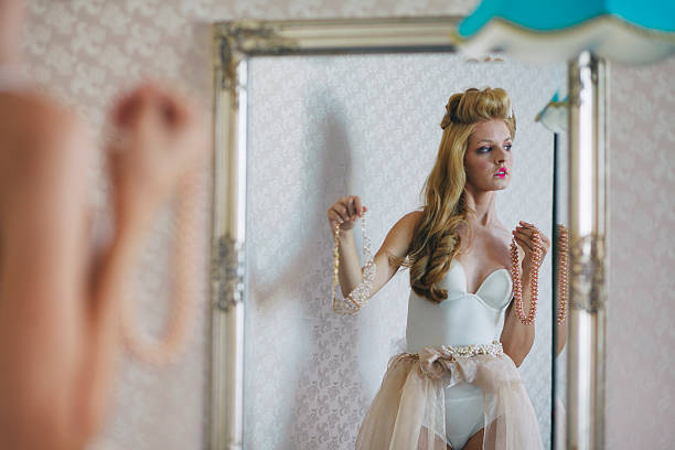 Elegant blonde bride in white strapless wedding gown getting dressed and applying pearl jewelry while looking in ornate vintage mirror. Soft natural lighting captures intimate bridal preparation moment in luxurious setting. Perfect for wedding planning, bridal fashion, luxury lifestyle and romantic celebration concepts.