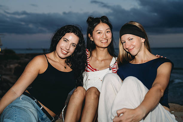 Three young women sitting together on the beach at dusk, smiling joyfully as they share laughter and create lasting memories