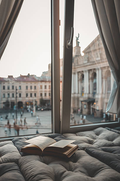 istock Cozy reading nook with an open book on a cushioned window seat overlooking a historic European city square and opera house at sunset through soft curtains 2231082633