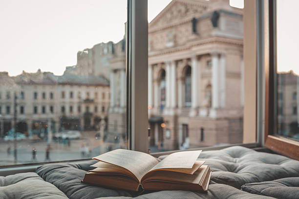 istock Cozy reading nook with an open book on a cushioned window seat overlooking a historic European city square and opera house at sunset through soft curtains 2231082636