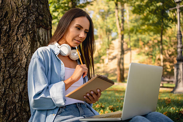 istock Woman sitting on grass at park working on laptop watching webinar writing notes. Female wearing earphones using laptop while sitting under a tree at park with bright sunlight from behind. 2231092070