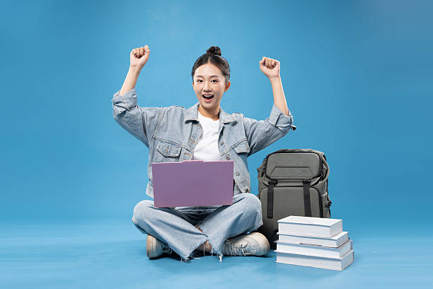 Enthusiastic student celebrates academic success with laptop and books on a blue background