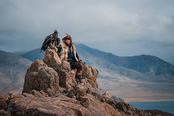 traditional Mongolian eagle hunter in fur clothing sits with his golden eagle on a rocky cliff, overlooking dramatic mountains and wide open sky. Mongolia.