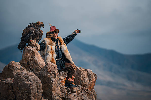 smiling Mongolian eagle hunter in traditional fur clothing sits on rocky terrain with a golden eagle perched on his arm, against a dramatic mountain backdrop. Mongolia.