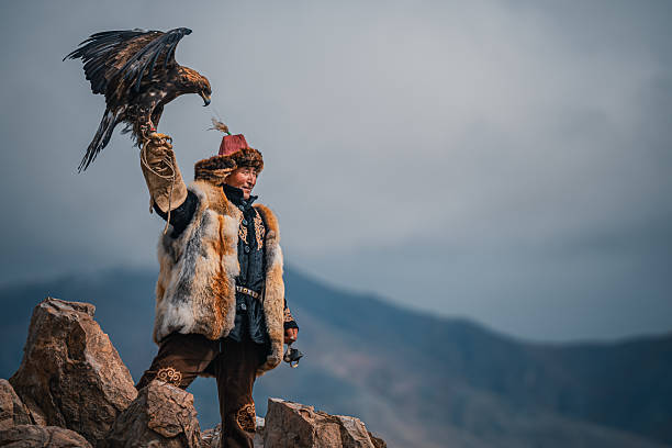 traditional Mongolian eagle hunter in fur clothing raises his golden eagle high while standing on rocky terrain against a mountain backdrop. Mongolia.