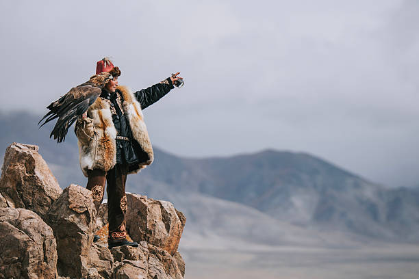 Mongolian eagle hunter in traditional fur clothing stands on rocky terrain with a golden eagle perched on his shoulder, pointing into the distance against a dramatic mountain backdrop. Mongolia.