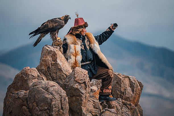 A traditional Kazakh eagle hunter in Mongolia poses on rocky terrain with a golden eagle perched on his arm, wearing fur clothing and looking into the distance. Mongolia.