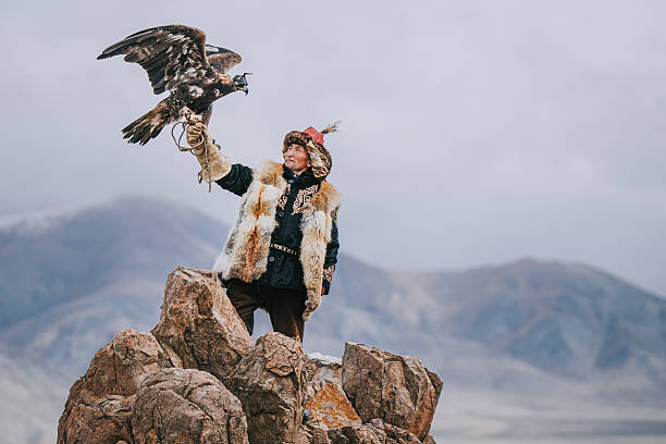 traditional Kazakh eagle hunter in Mongolia stands proudly on rocky terrain, holding a golden eagle against a dramatic mountain backdrop. This image represents cultural heritage, strength, and the ancient tradition of falconry. Mongolia.