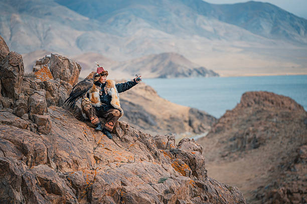 traditional Mongolian eagle hunter in fur clothing sits on rocky terrain with his golden eagle, overlooking a lake and mountain landscape. Mongolia.