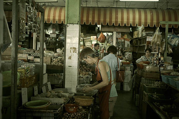A wide-angle, documentary-style shot showing the bustling interior of a traditional market. The image focuses on a stall owner in the foreground attending to produce, while other vendors and a customer are visible in the background, surrounded by various goods under a striped awning. This photo captures the authentic and lively atmosphere of a local market in Kuala Lumpur.