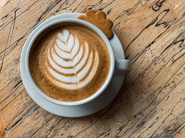 Close-up overhead shot of a cup of coffee on a saucer on a wooden surface with intricate grain. The coffee features detailed latte art and there is a ginger biscuit on the saucer.
