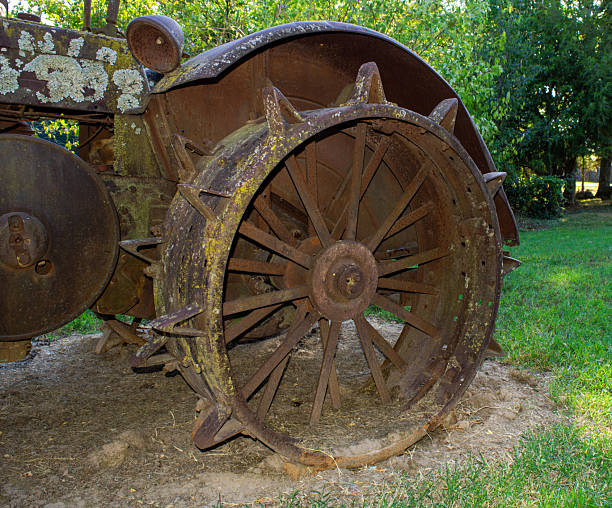 Close up look of a weathered vintage tractor wheel made of heavy cast iron displayed on a green lawn with slightly defocused green trees in the background.