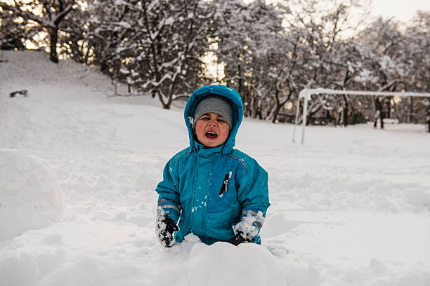 Young boy wearing blue snowsuit playing in deep snow during cold winter day, shivering and looking uncomfortable