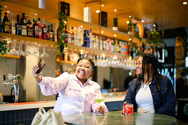 istock Smiling lesbian couple sitting at an illuminated bar, taking a selfie together while enjoying colourful drinks 2231131347