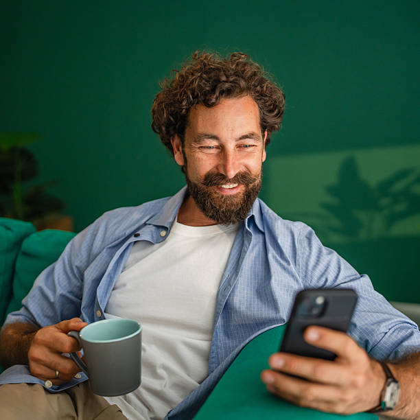 istock Smiling man using smart phone and drinking coffee on sofa at home 2231135401