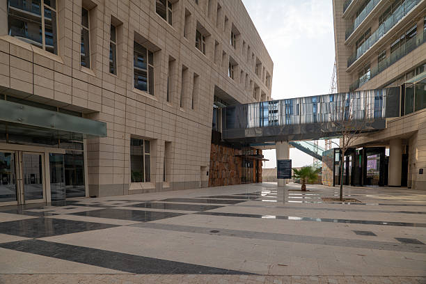 Modern Buildings with Skybridge in King Abdullah Financial District, Riyadh. A shot of two modern office buildings connected by a sleek, elevated skybridge in Riyadh's King Abdullah Financial District (KAFD). The buildings have a clean, contemporary design with a stone-like facade and large windows. The ground is covered in a striking black and white tiled pattern, adding a unique geometric element to the scene. High quality photo