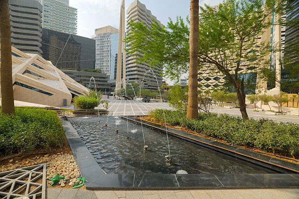 Fountain and Public Plaza in Boulevard, Riyadh. A vibrant shot of a modern public plaza in Boulevard. In the foreground, a sleek, black-tiled fountain with multiple jets creates a cooling effect, surrounded by lush green shrubs and trees. The background features various contemporary skyscrapers with unique architectural designs, including one with a distinctive geometric facade. High quality photo