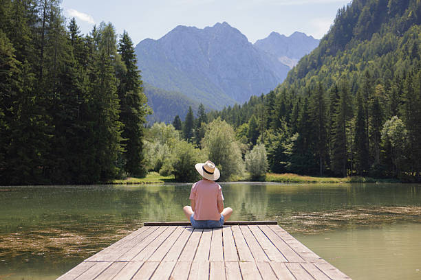 A person sits peacefully on a wooden dock, gazing at the tranquil lake surrounded by lush trees and majestic mountains on a sunny day.