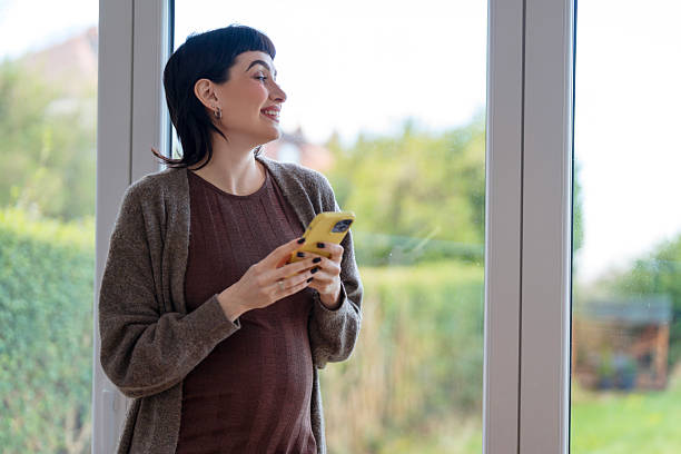 istock Pregnant woman standing by a large window, smiling while using her smartphone in a bright and cozy indoor space in the afternoon 2231155015