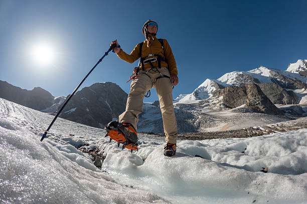Female climber wearing helmet and sunglasses hiking on icy glacier terrain with trekking pole. Adventurous woman trekking in snowy mountain landscape. Outdoor mountaineering and alpine exploration.