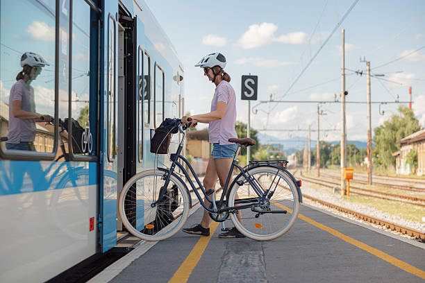 A person in shorts and a helmet is loading a bicycle onto a train at a busy station on a sunny day, ready to start their travel.