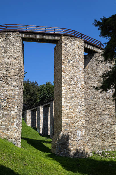 ancient stone bridge with a wooden walkway on top, connecting two tall stone columns, against a clear blue sky, with green grass in front