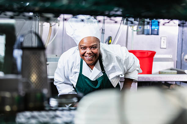 A mature black woman in a commercial kitchen wearing chef's whites, bending as she smiles at the camera, looking through the shelves of a wire storage unit.