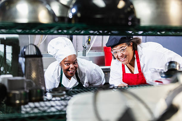 Two black women working as cooks in a restaurant standing in the kitchen, smiling and looking at the camera through the shelves of a wire storage unit.