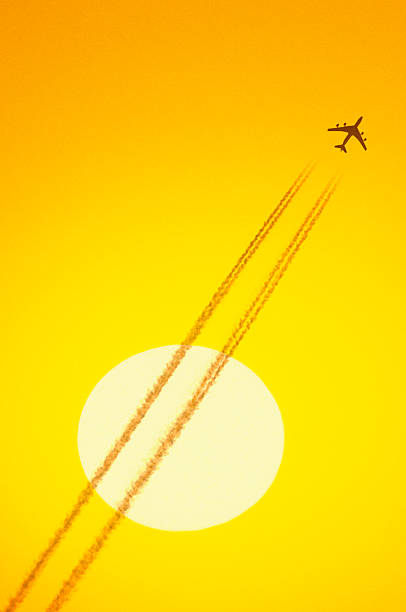 A silhouetted jet, vapor trails and a big sun in an orange sky.