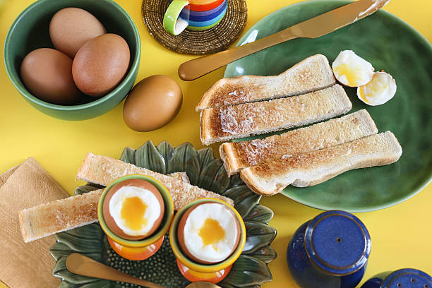 Stock photo showing close-up, elevated view of a breakfast scene on a yellow surface. The scene is set with a variety of crockery including dark blue salt and pepper shakers, a dark green, sunflower shaped plate with two, rainbow coloured, striped egg cups containing oozing, soft boiled eggs with a metal teaspoon, a green tea plate of buttered white toasted bread soldiers with a knife, as well as a dark green bowl full of chickens eggs and a black, espresso coffee in a yellow, orange, red, indigo, violet, turquoise blue and light green striped espresso cup on a coaster.