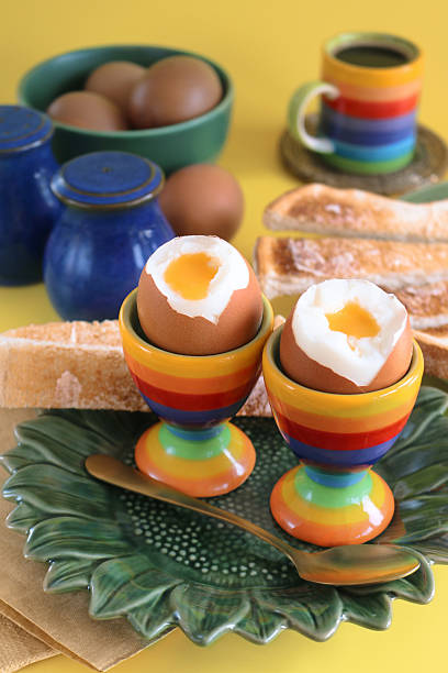 Stock photo showing close-up view of a breakfast scene on a yellow surface. The scene is set with a variety of crockery including dark blue salt and pepper shakers, a dark green, sunflower shaped plate with two, rainbow coloured, striped egg cups containing oozing, soft boiled eggs with a metal teaspoon, a green tea plate of buttered white toasted bread soldiers, as well as a dark green bowl full of chickens eggs and a black, espresso coffee in a yellow, orange, red, indigo, violet, turquoise blue and light green striped espresso cup on a coaster.