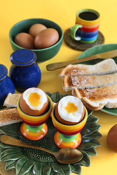 istock Image of breakfast scene, green sunflower plate, two egg cups, oozing soft boiled eggs, white buttered toasted bread soldiers, metal teaspoon and knife, blue salt and pepper pots, bowl of eggs, rainbow striped espresso cup, yellow background, elevated vie 2231195060