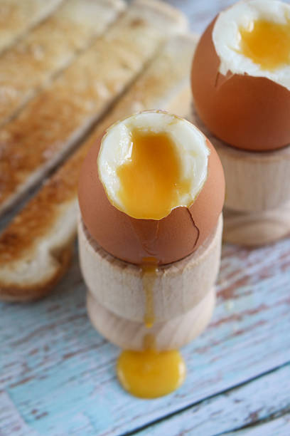 istock Full frame image of white buttered toasted bread slices, two soft boiled egg with runny yellow yolks in wooden egg cups, white buttered toasted bread soldiers, whitewashed wood grain background, elevated view, focus on foreground 2231197895