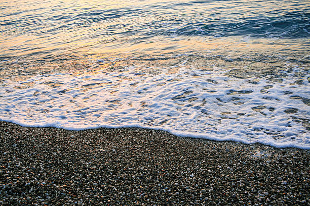 Gentle waves are lapping against a pebbled shore, creating a tranquil coastal scene during sunset, with soft colors reflecting on the water's surface and a peaceful ambiance surrounding the area
