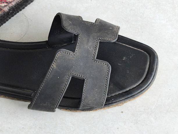 A single black leather sandal, featuring a unique H-shaped strap design, rests on a subtly textured light-colored background.