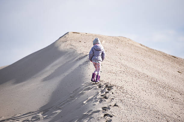 Child walking up a sandy hill in winter clothes, leaving footprints in the sand on a bright day