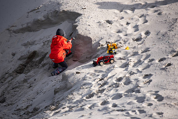 Young child wearing a red coat playing with toy trucks in a sandpit, building and digging on a sunny day