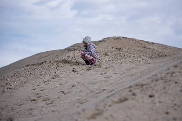Girl crouching on a sand dune, feeling the texture of the sand while surrounded by a cloudy sky, embracing the beauty of nature