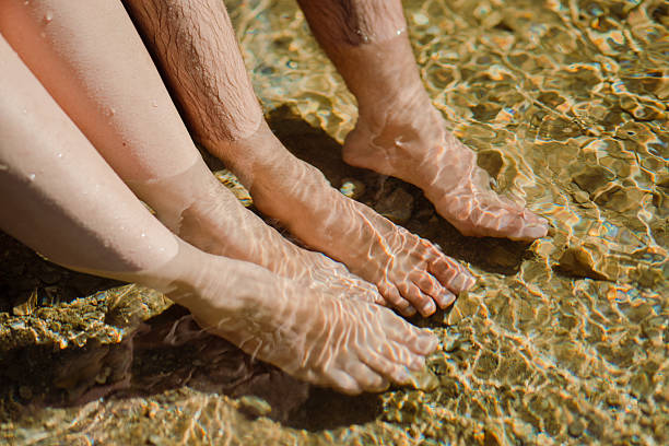 Close-up view of a man and woman relaxing with their feet submerged in a shallow, clear stream. The sunlight creates beautiful reflections on the water, capturing a peaceful and intimate summer moment. Perfect representation of relaxation, connection, and nature.