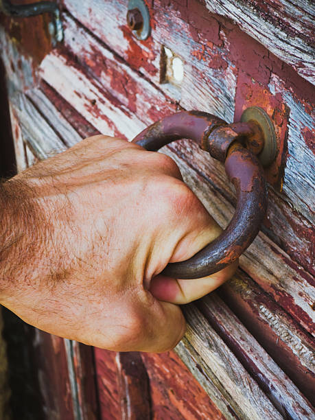 Close-up of a human hand gripping and pulling a rusty metal handle on a weathered wooden door with peeling paint. The image symbolizes strength, opening, access, history, and rustic vintage character.