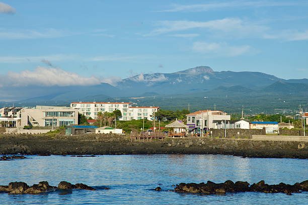 A view over Gwideok Village shows low seaside buildings and a waterfront pavilion with broad Hallasan rising inland, its slopes partially veiled by morning clouds above the coast.