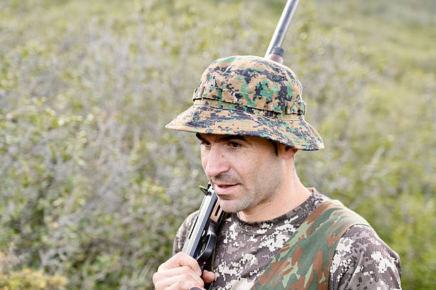 A man in camouflage hat and shirt holding a gun. The man is looking at the camera. Concept of adventure and excitement, as the man is preparing for a hunting trip