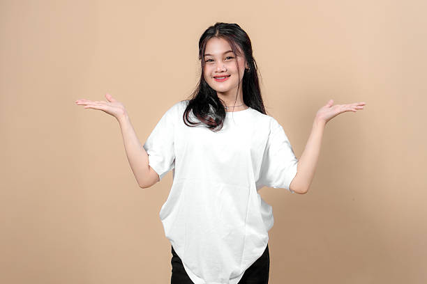 istock Young Asian woman in a plain white oversized t-shirt smiling and posing with open hands against a beige background, presenting or comparing something with cheerful expression. 2231281462