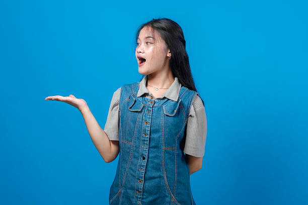 istock Surprised young Asian woman looking sideways and presenting something on her open palm, wearing denim dress and gray shirt, standing confidently on bright blue studio background. 2231282067