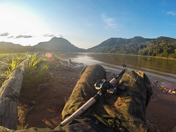 Fisherman rests on the riverbank with his fishing rod, enjoying the tranquil sunset over the amazon rainforest