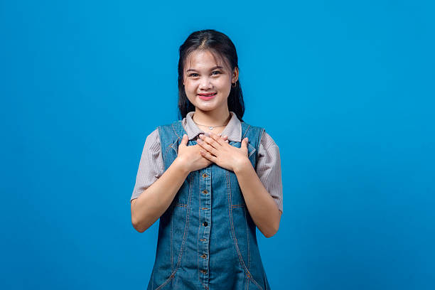 istock Young Asian woman smiling gently with both hands on her chest, expressing gratitude, sincerity, or appreciation, standing against a plain blue background. Honest and heartfelt emotion. 2231283974