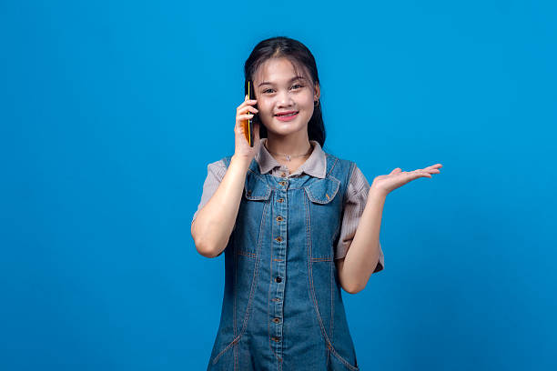 istock Young Asian woman smiling while talking on a smartphone and raising one hand as if presenting something, wearing a denim dress, isolated on blue background. Friendly and inviting gesture. 2231284611