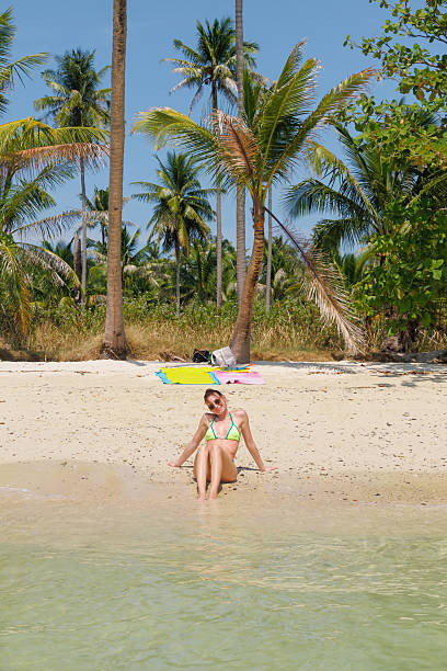 A woman relaxes on the sandy shore of a tropical beach in Thailand. Surrounded by palm trees and clear blue water, she enjoys the warm sun.